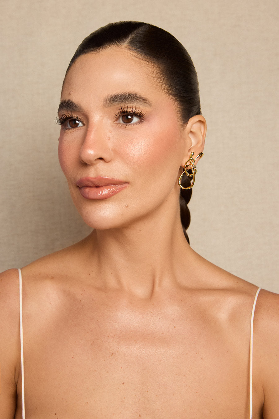 Um retrato em close de uma mulher de cabelos escuros presos para trás, usando brincos de argola dourados com um design circular texturizado e em camadas. Ela veste uma blusa branca de alças finas, tendo ao fundo uma superfície bege neutra e texturizada.