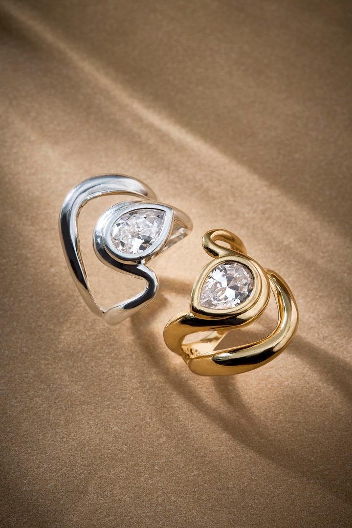 Anel Audrey regulável em dourado e prateado, com curvas suaves e cristal central. Uma joia delicada e contemporânea que traduz charme, elegância e refinamento.