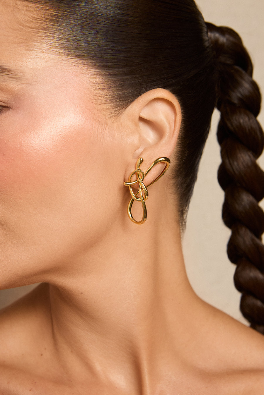 Um close-up de perfil lateral de uma mulher usando um brinco dourado em formato de laço abstrato, com o cabelo escuro preso em uma trança, contra um fundo bege neutro.
