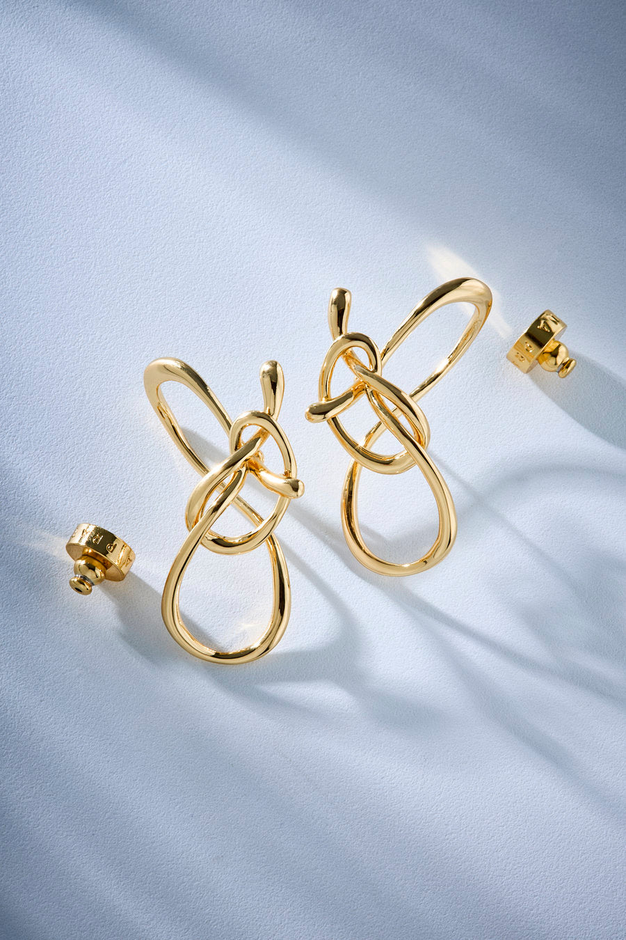 Brincos de destaque em tom dourado com design de laços entrelaçados e abstratos, exibidos sobre uma superfície macia e branca com aspecto de cetim, acompanhados por algumas peças pequenas e coordenadas nas proximidades.
