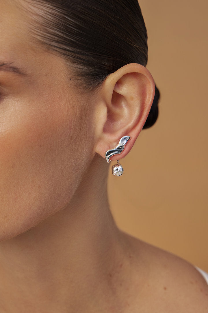 Brinco Frida em tom prateado, com curvas orgânicas e detalhe esférico em uma composição escultural. Uma joia contemporânea que traduz expressão, personalidade e força feminina.