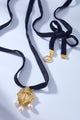 Choker de veludo preto com pingente dourado em formato de escudo, detalhe central em relevo e três pérolas pendentes.