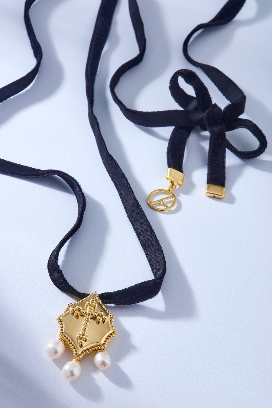 Choker de veludo preto com pingente dourado em formato de escudo, detalhe central em relevo e três pérolas pendentes.