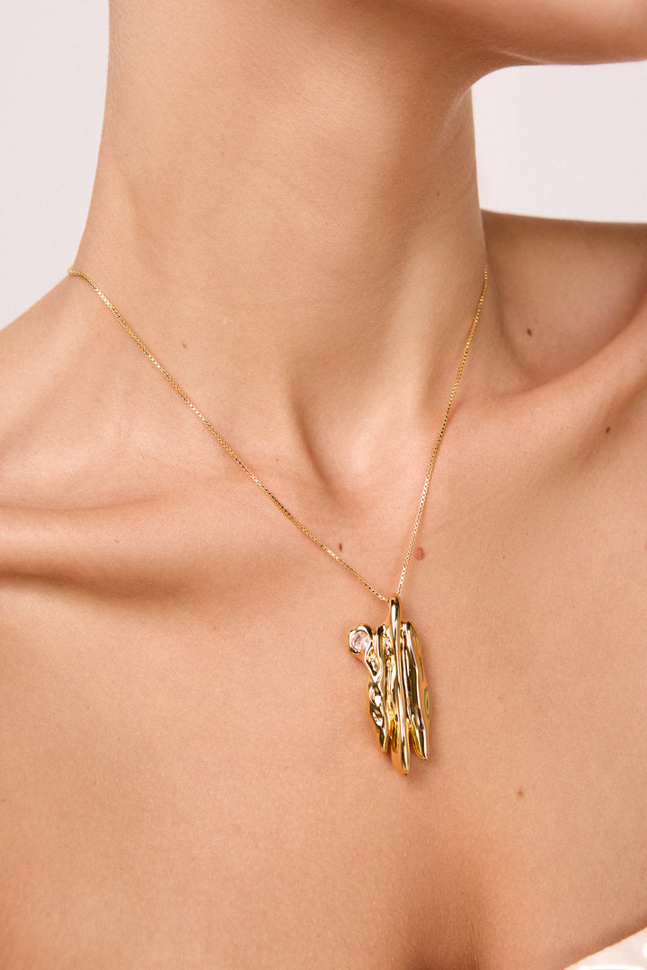 Colar Temporae dourado com corrente delicada e pingente escultural texturizado, adornado com zircônia, sobre fundo claro.