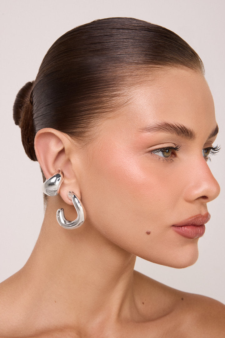 Piercing Marea prateado em formato de gota orgânica assimétrica, com volume tridimensional e acabamento espelhado sobre fundo claro.