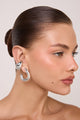 Piercing Marea prateado em formato de gota orgânica assimétrica, com volume tridimensional e acabamento espelhado sobre fundo claro.