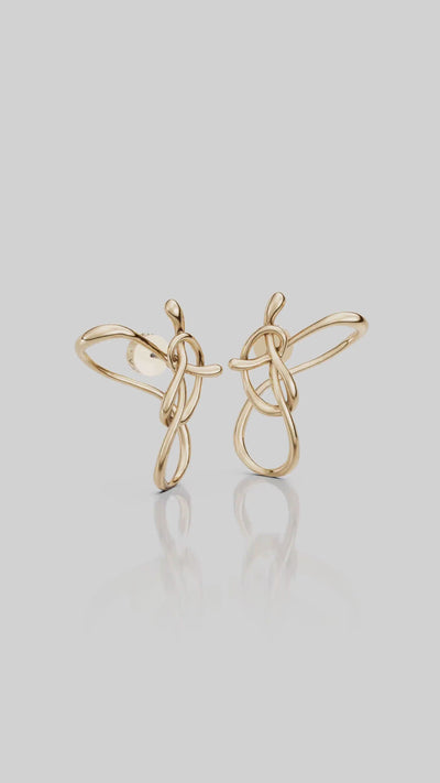  Um close-up da orelha de uma modelo usando um brinco de dourado com um design de nó abstrato entrelaçado e um pingente em formato de gota.
